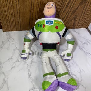 May include: A plush toy of Buzz Lightyear from the Toy Story franchise. The toy is sitting with its legs crossed and has a purple, white, and green space suit. The toy has a large head with a smiling face and a purple helmet. The toy's chest has a green patch with the words "SPACE RANGER" and "LIGHTYEAR" on it.