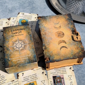 May include: A wooden box with a lock and a distressed, aged finish. The box has a design of three moons on the front. The box is sitting on a table with a deck of tarot cards. The cards have a vintage design and feature a sun with a face on the front. The text on the cards reads "Learning Tarot by TaroTerra".