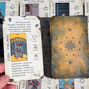 May include: A tarot card with the text "Cups - Feelings" and the number "9" is held next to a deck of cards. The card features an illustration of a person and cups. The back of the deck has a starburst design.