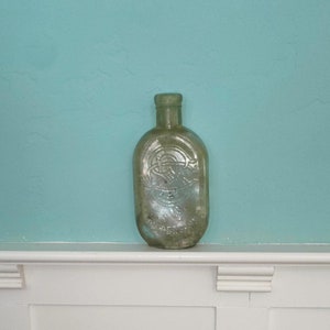 Può includere: Una bottiglia di vetro trasparente con una superficie testurizzata e un'etichetta che recita "TRADE MARK REG. U.S. PAT. OFF.". La bottiglia è appoggiata su uno scaffale bianco con boiserie.