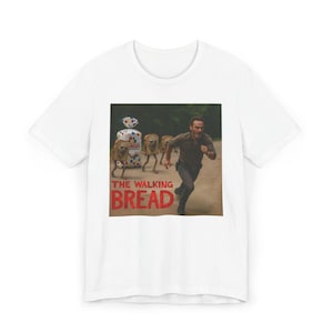Puede incluir: Camiseta blanca con un gráfico humorístico. El gráfico muestra a un hombre corriendo de barras de pan con caras, con el texto "THE WALKING BREAD" en letras rojas. La imagen es una parodia de un programa de televisión popular.