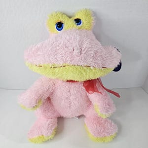 May include: A plush frog toy in shades of pink and yellow, with large blue eyes and a red ribbon tied around its neck. The frog has a soft, fuzzy texture and a stitched mouth, giving it a friendly appearance. A cute stuffed animal.