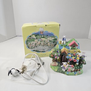 May include: A decorative porcelain house with a green roof, colorful flowers, and bunny figurines. The house has a sign that says "Welcome" and "My Garden". A power cord with a bulb is included. The box has a picture of a village.