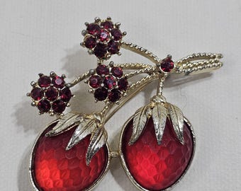 May include: Vintage brooch featuring a gold-tone metal design of a branch with two red strawberry-shaped pendants and three clusters of red rhinestones. The brooch has a textured finish and a pin closure on the back.