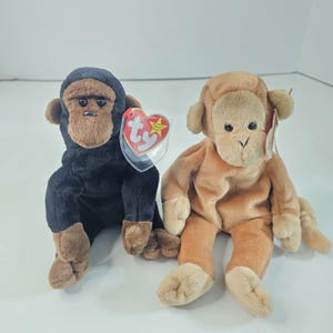 May include: Two plush monkey toys. One is black with brown accents, the other is tan. Both have the Ty heart-shaped tag attached. The monkeys are sitting on a white surface.