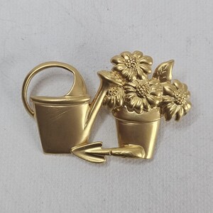 May include: A gold-colored brooch featuring a watering can, a trowel, and a pot of flowers. The flowers have detailed petals and leaves. The brooch has a matte finish and is likely made of metal.