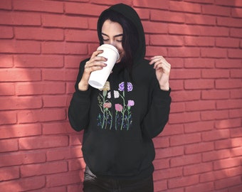 Peut inclure: Un sweat à capuche noir avec un motif floral composé de œillets roses, violets et blancs. Le sweat à capuche a une capuche à cordon et une poche kangourou.