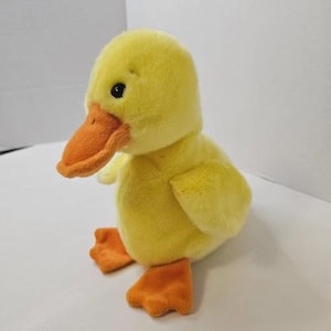 May include: A plush yellow duckling toy with soft fur, featuring a bright orange beak and matching orange webbed feet. The duckling has small black eyes and is positioned in a standing pose, showcasing a rounded body and small wings.