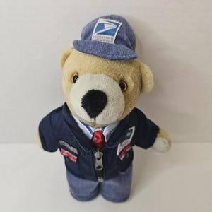 May include: A plush teddy bear wearing a blue United States Postal Service cap with the eagle logo, a navy blue zip-up jacket with matching patches, a light blue collared shirt, and a red necktie. The bear has tan fur and a black nose.