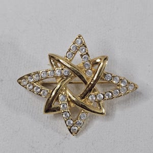 May include: Gold-tone starburst brooch with clear crystal accents. The brooch features a four-pointed star design with interwoven elements, each point outlined with sparkling crystals. A classic accessory.