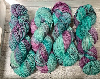 May include: Four skeins of hand-dyed yarn in a wooden tray. The yarn is a blend of teal, purple, and pink with black speckles. The yarn is arranged in a twisted, bulky form, ideal for knitting or crocheting projects.
