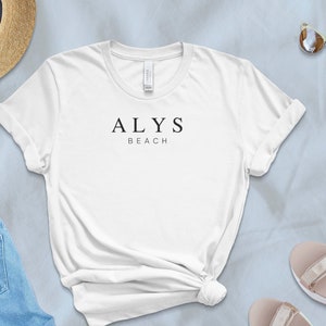May include: White t-shirt with the text "ALYS BEACH" printed on the front. The shirt is laid out on a light blue surface with a straw hat, denim shorts, and sandals.