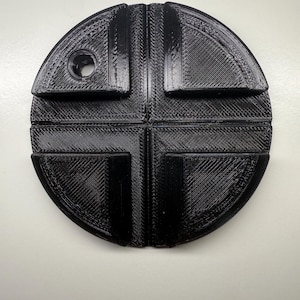 Può includere: Un oggetto nero, circolare, stampato in 3D con un design a forma di croce. L'oggetto ha una superficie strutturata e un foro su un lato. Il design è diviso in quattro quadranti, ciascuno con un bordo curvo.