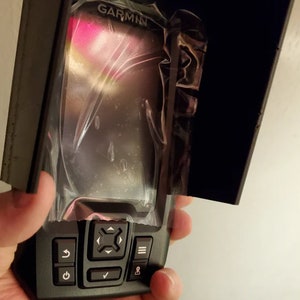 May include: A black Garmin device with a protective cover. The device has a screen and several buttons, including power, navigation, and menu options. The Garmin logo is visible at the top. The device is held in a hand.