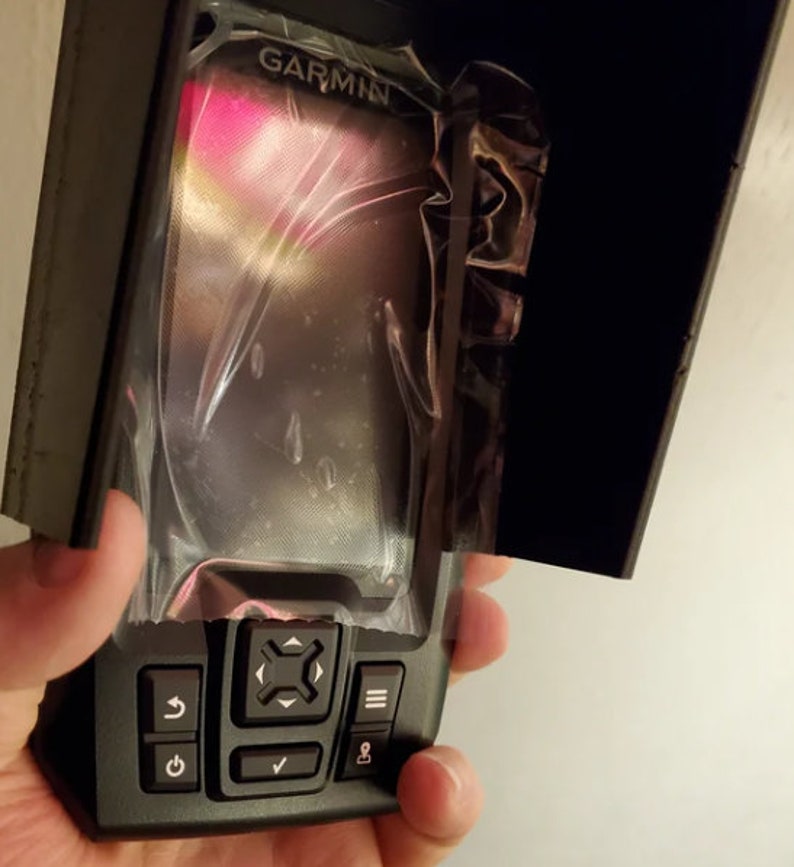 May include: A black Garmin device with a protective cover. The device has a screen and several buttons, including power, navigation, and menu options. The Garmin logo is visible at the top. The device is held in a hand.