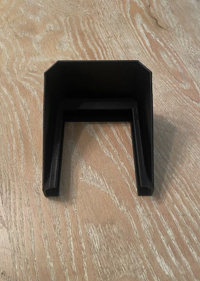 May include: A black, rectangular, 3D-printed object with a U-shaped opening. The object has a flat top and two vertical sides, and it is sitting on a light-coloured wooden surface.