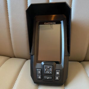 May include: A black Garmin fish finder with a protective visor. The device has a rectangular screen and several control buttons below the screen. The word "GARMIN" is printed above the screen.