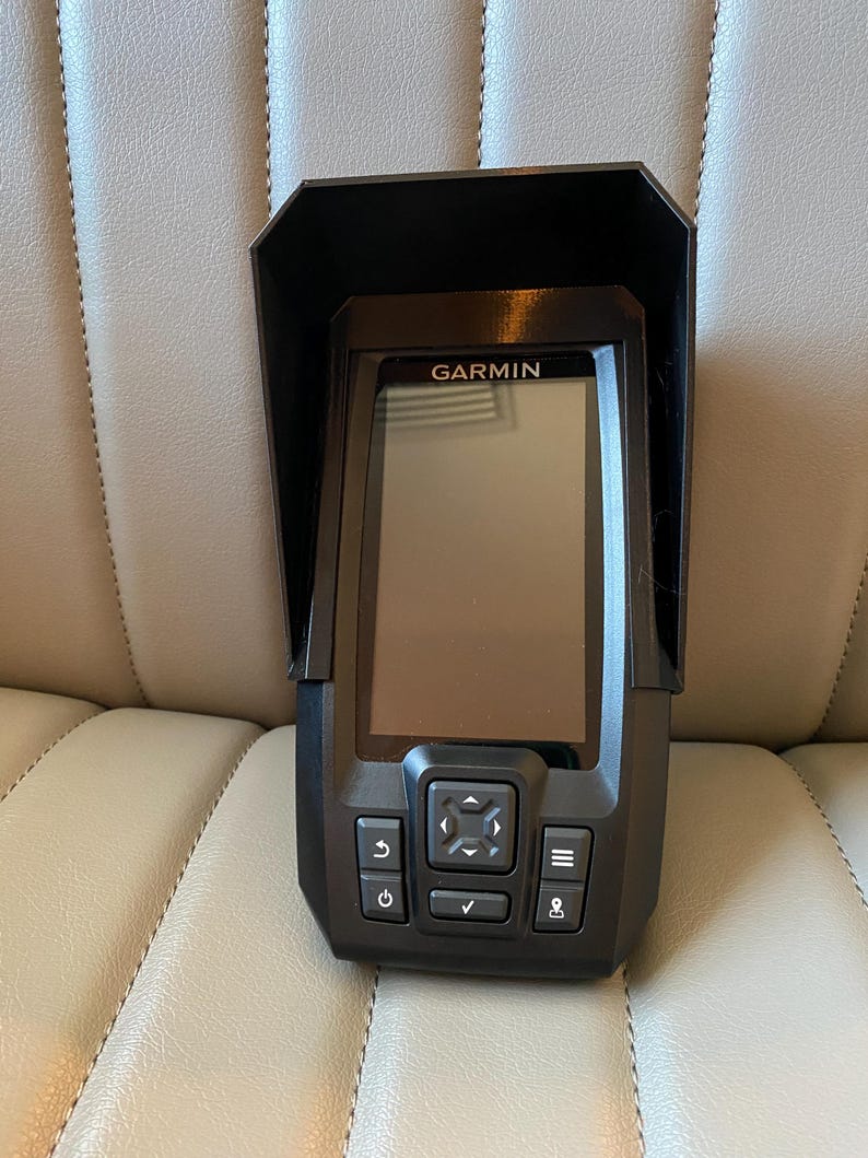 May include: A black Garmin fish finder with a protective visor. The device has a rectangular screen and several control buttons below the screen. The word "GARMIN" is printed above the screen.