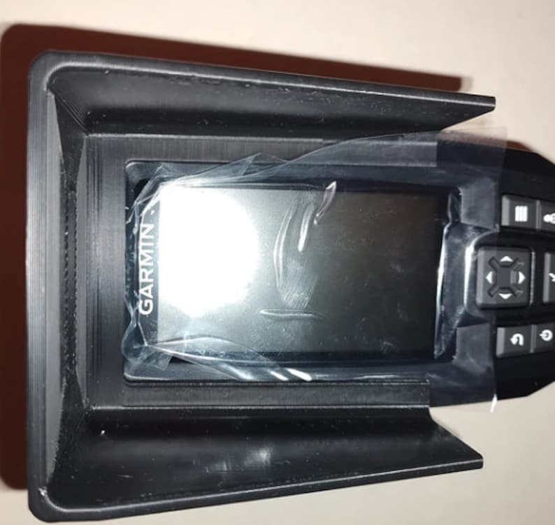 May include: A black Garmin device with a screen covered in plastic wrap, set in a black holder. The device has several buttons on the right side. The Garmin logo is visible on the screen.