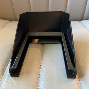 May include: A black plastic, U-shaped object with a textured interior. The object has a rectangular top and two angled legs. The legs are connected by a horizontal bar. The object is sitting on a light-coloured surface.