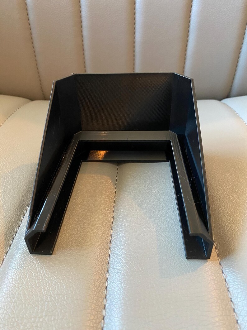 May include: A black plastic, U-shaped object with a textured interior. The object has a rectangular top and two angled legs. The legs are connected by a horizontal bar. The object is sitting on a light-coloured surface.