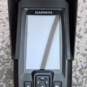 May include: Black Garmin fish finder with a protective visor. The device has a screen and several buttons. The word "GARMIN" is printed on the top of the device.