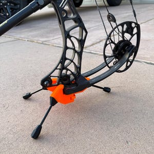 May include: A black compound bow resting on a bright orange tripod stand. The bow features a complex skeletonized riser design, a circular cam system with black cables, and limbs labeled with Switchweight technology. The stand has three thin legs.