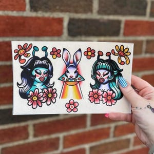 May include: A rectangular print featuring three central illustrations: a blue-skinned figure with antennae, a rabbit inside a flying saucer with a rainbow beam, and a green-haired figure with antennae, all surrounded by small orange and pink flowers.