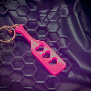 May include: A pink paddle keychain with three heart cutouts. The keychain is on a black honeycomb-patterned surface.