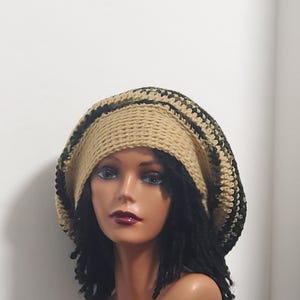 May include: A tan and black crocheted hat with a wide brim. The hat is worn by a mannequin with black dreadlocks. The hat's design features a tan base with black and olive green stripes around the brim.