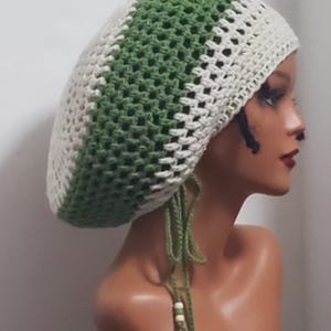 May include: A crocheted tam hat featuring alternating vertical stripes of cream and sage green yarn. The hat has a loose, textured stitch pattern and includes two thin green ties hanging from the side, each finished with small white beads.