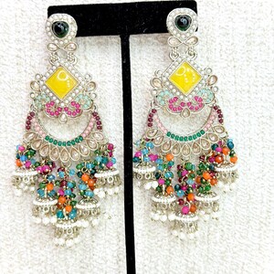 May include: Silver-toned chandelier earrings with a yellow diamond-shaped center and colorful bead accents. The earrings feature a green stone at the top and small white bell-shaped dangles at the bottom. The earrings are displayed on a black stand.