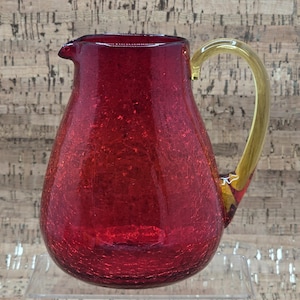 Könnte beinhalten: Ein roter Glaskrug mit Craquelé-Finish. Der Krug hat einen abgerundeten Körper, einen Ausguss und einen goldfarbenen Griff. Der Krug steht auf einer transparenten Glasoberfläche.