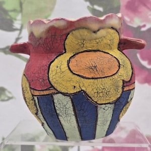 Könnte beinhalten: Handbemalter Keramiktopf mit einem skurrilen Design. Der Topf hat eine rote Oberseite mit einem gewellten Rand, eine große gelbe Blume und blau-weiß gestreifte Abschnitte. Kleine Griffe ragen von den Seiten heraus.