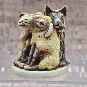 May include: A detailed figurine depicting three animals: two sheep and a dog. The sheep are light beige with textured wool, while the dog is gray and brown. The animals are huddled together on a white base, creating a heartwarming scene.