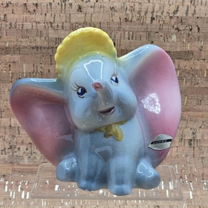 May include: A ceramic figurine of Dumbo the elephant, featuring a light blue body, large pink and blue ears, and a yellow hat. The figurine has a smiling expression and a small red nose. A sticker with the word "DUMBO" is on the side.