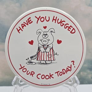 以下が含まれることがあります： 赤い縁取りと「Have You Hugged Your Cook Today?」の文字が入った丸い白いボタン。ストライプのエプロンを着てスプーンを持った漫画の豚が描かれています。赤いハートが散りばめられています。