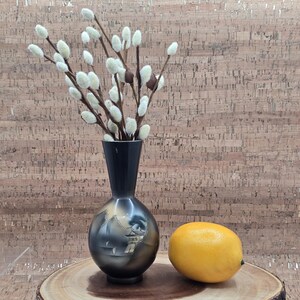 Puede incluir: Un jarrón negro y dorado con un diseño de paisaje contiene ramas de sauce blanco y marrón. Un limón amarillo se encuentra en una tabla redonda de madera. La tabla está sobre un soporte transparente.