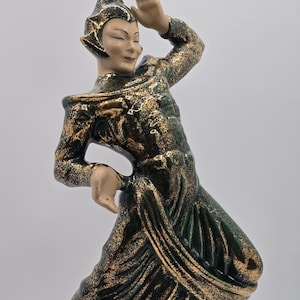 May include: A ceramic figurine of a dancer wearing a green and gold costume. The dancer is posed in a dynamic, flowing movement, with one arm raised and the other extended to the side. The figurine is detailed with intricate patterns and textures.
