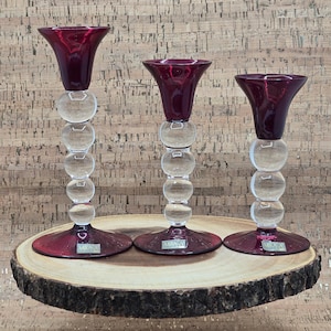 Puede incluir: Tres candelabros decorativos. Cada soporte tiene una parte superior acampanada de color rojo intenso y un tallo transparente y esférico. Las bases también son de color rojo intenso. Los candelabros se exhiben sobre una losa de madera redonda con borde natural.