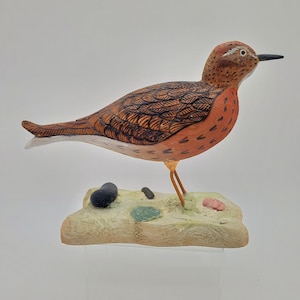 May include: Hand-carved wooden bird sculpture of a shorebird with brown and orange feathers standing on a wooden base with green and brown accents.