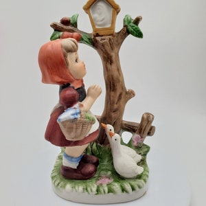 May include: A porcelain figurine of a girl in a red hooded dress holding a basket, standing next to a tree with a birdhouse and two white geese.