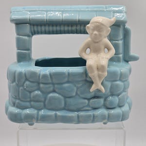 Könnte beinhalten: Ein hellblauer Keramik-Pflanztopf in Form eines Steinbrunnens mit einem weißen Keramik-Zwerg, der auf dem Rand sitzt. Der Topf hat eine rechteckige Öffnung zum Pflanzen.