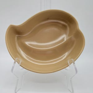 May include: A light brown ceramic divided dish with a unique curved shape. The dish is on a clear acrylic stand.
