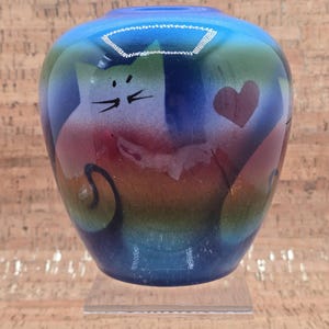 May include: A colourful, hand-painted ceramic vase with a cat design. The vase features a gradient of blues, greens, and reds, with a cat face and a heart. The vase is approximately 13 cm tall and 10 cm wide.