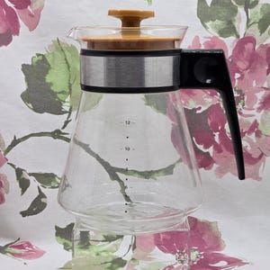 May include: A clear glass coffee carafe with a brown lid and black handle. The carafe has a silver band around the middle and measurement markings from 6 to 12. The carafe is sitting on a clear stand.