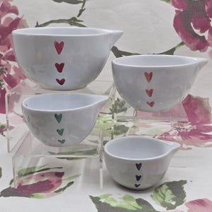 Può includere: Set di quattro ciotole dosatrici in ceramica bianca con beccucci. Ogni ciotola è decorata con una serie di tre cuori rossi, arancioni, verdi e viola. Le ciotole sono esposte su supporti in acrilico trasparente.