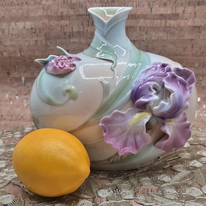 Puede incluir: Un jarrón de porcelana con un cuerpo liso de color crema, decorado con suaves acentos de hojas verdes y flores de iris moradas esculpidas en tres dimensiones. Un limón amarillo brillante se sitúa en primer plano, dando una escala clara.