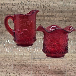 Puede incluir: Dos piezas de servicio de vidrio rojo rubí. Una jarra con asa y boquilla, y un cuenco con borde festoneado. Ambas presentan un patrón en relieve de hojas y bayas. Se exhiben sobre soportes de acrílico transparente.