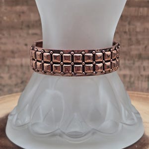 Pode incluir: Uma pulseira cor de cobre com um design geométrico. A pulseira apresenta duas fileiras de formas quadradas, cada uma com um pequeno ponto no centro. A pulseira é exibida em uma forma de vidro fosco.